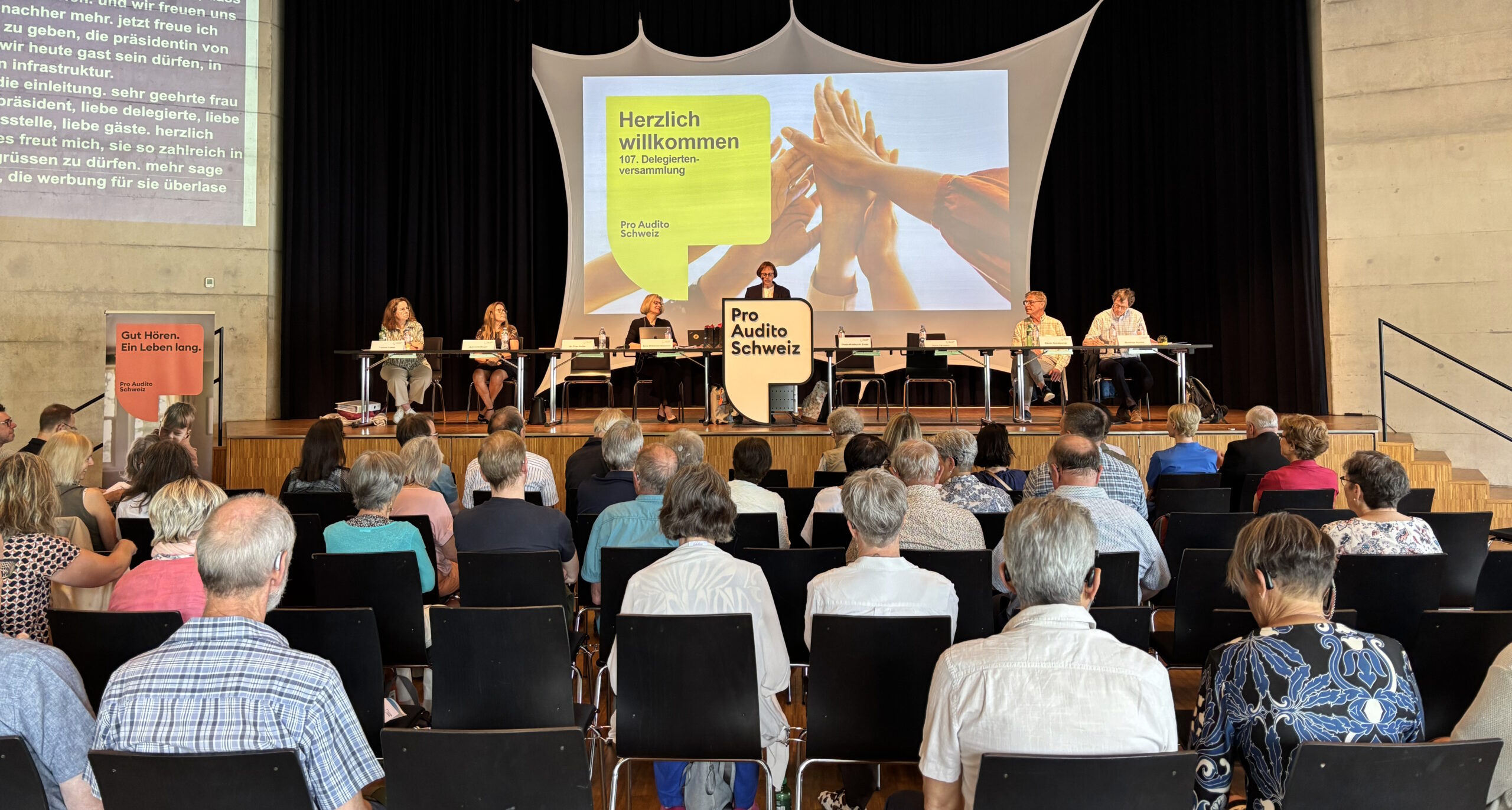 Ansicht der Bühne der Delegiertenversammlung 2026 in Winterthur. Die Rednerin Danielle Müller steht am Pult, wähend der Zentralvorstand und die Zuschauer:innen die Rede verfolgen
